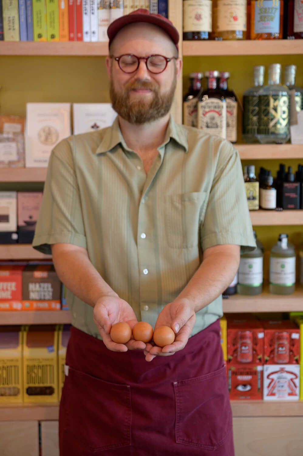 Bearded man wearing glasses and apron holds fresh brown eggs in a shop with books and bottles on shelves.