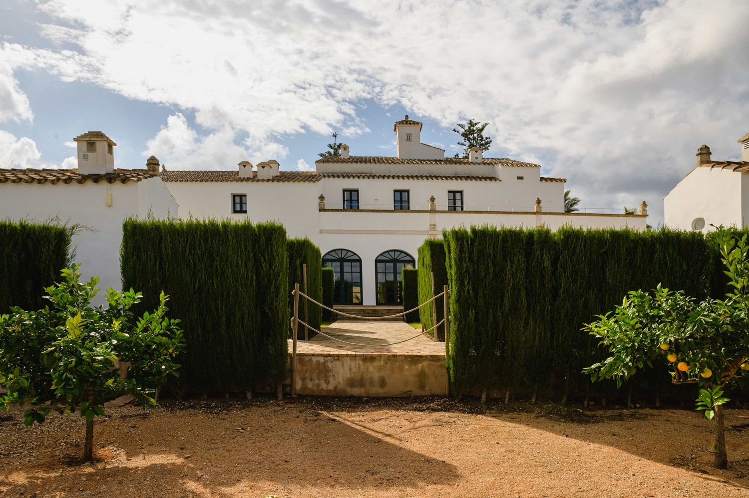 White Spanish villa with cypress hedges and citrus trees lining a gravel path under a cloudy sky.