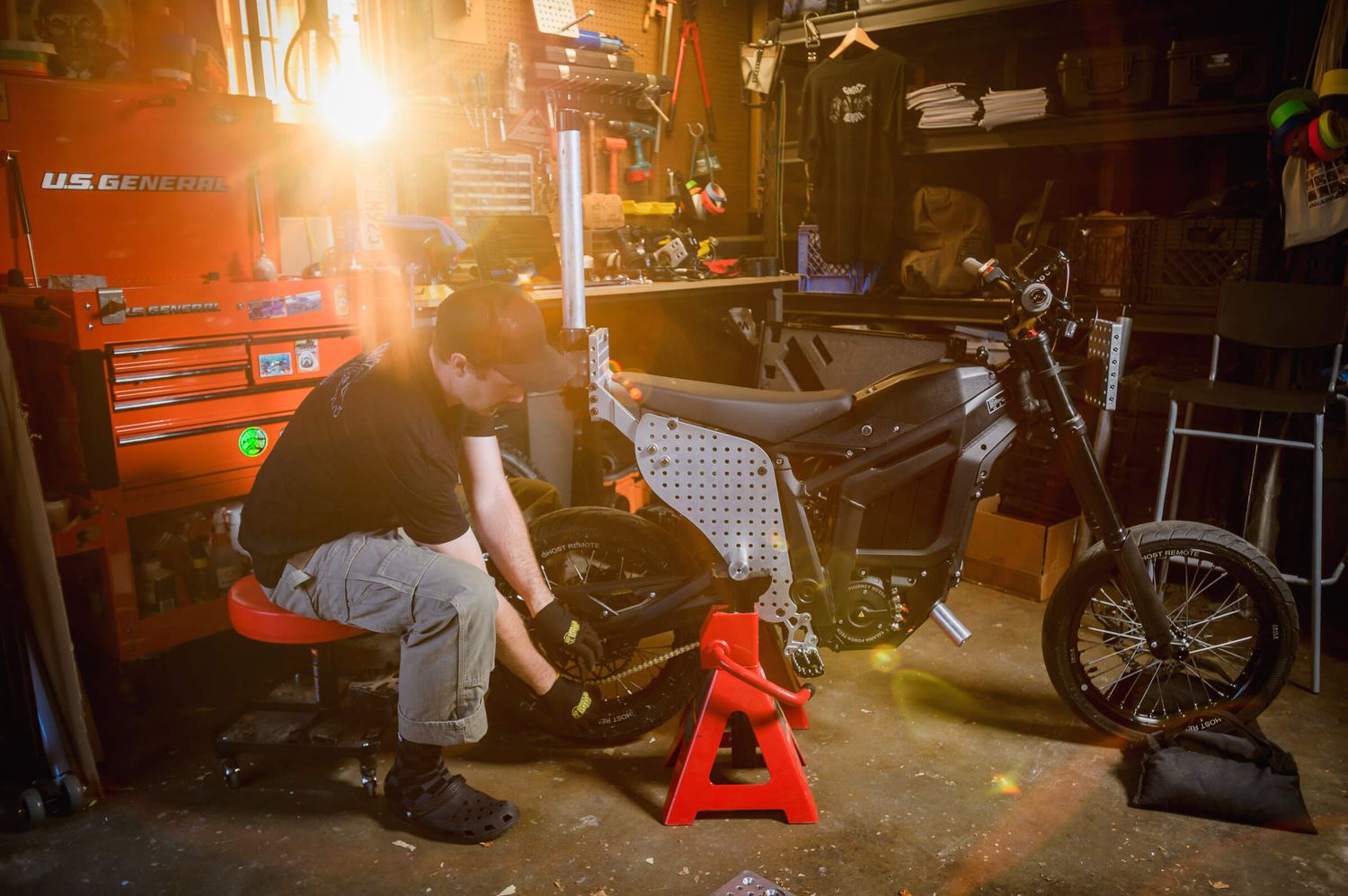 Mechanic working on a motorcycle in a garage workshop with red jack stands and tool chest.