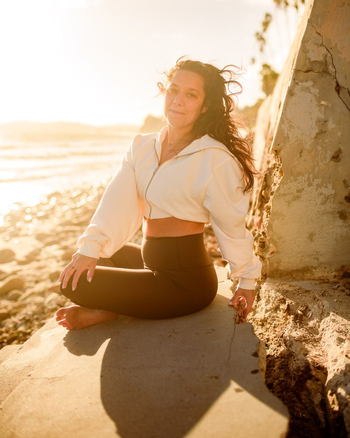 Woman in white crop hoodie and black leggings sitting cross-legged on rocks at golden hour beach.