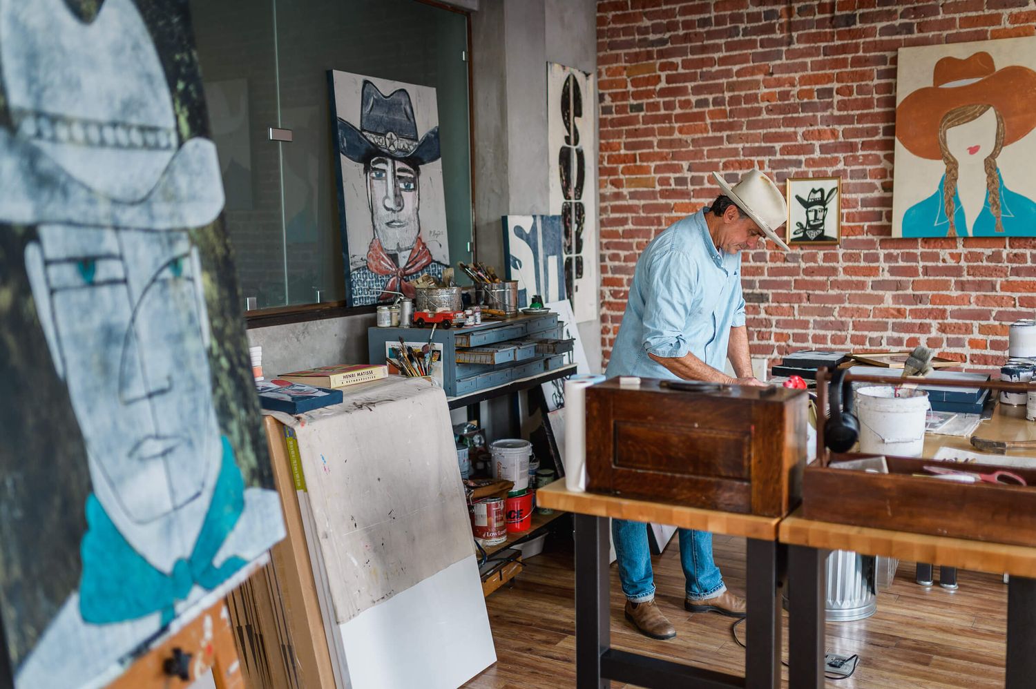 Artist working in a studio surrounded by large portrait paintings and art supplies in a brick-walled gallery space.