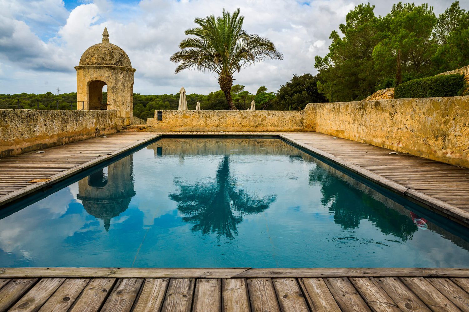 Historic stone pool with wooden deck reflecting a tall palm tree and ornate turret under a cloudy sky.