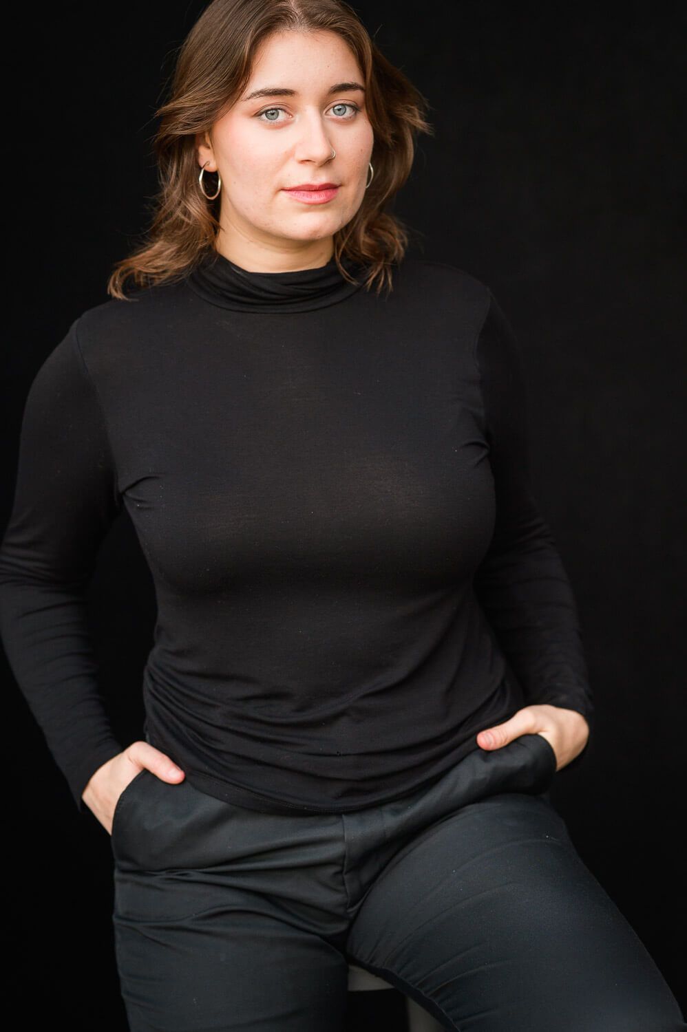 Woman in black turtleneck and dark pants poses confidently against a dark background with hands in pockets.