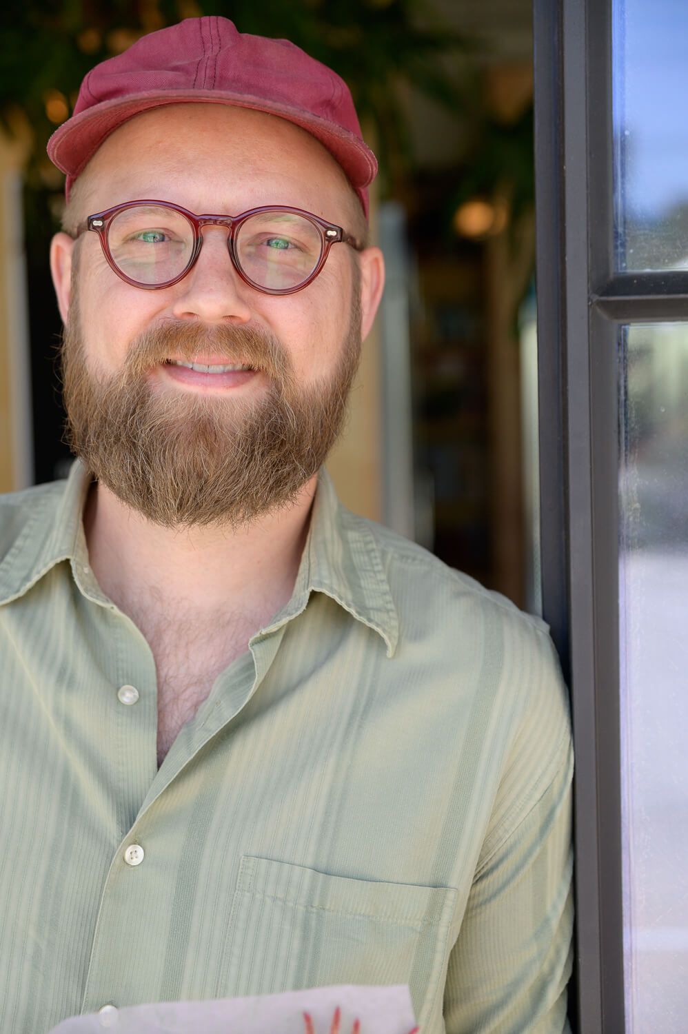 Smiling bearded man wearing round glasses and a red cap, dressed in a green striped shirt, standing near a doorway.
