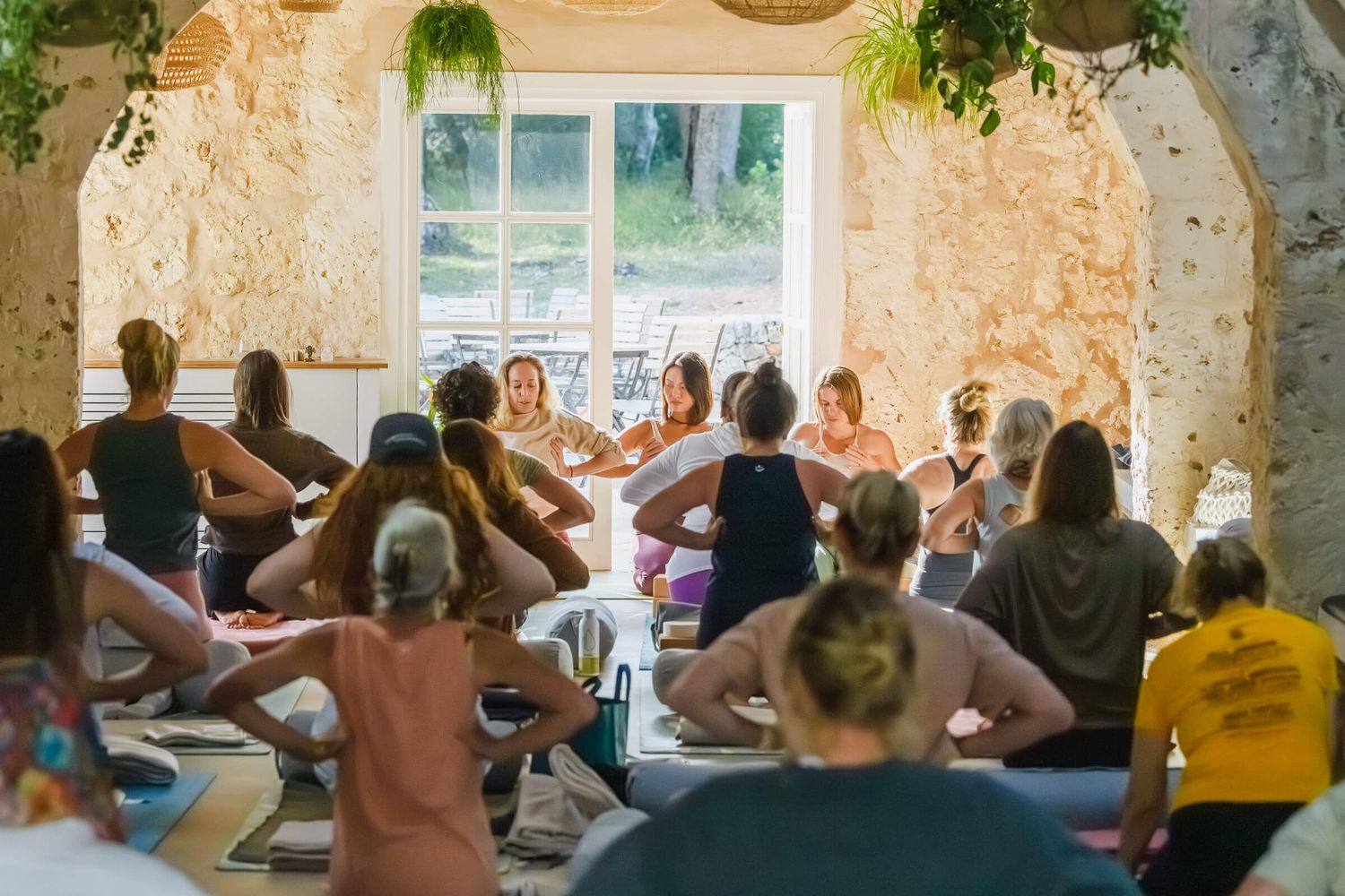 Large yoga retreat group session in rustic stone room with natural light from wide window.