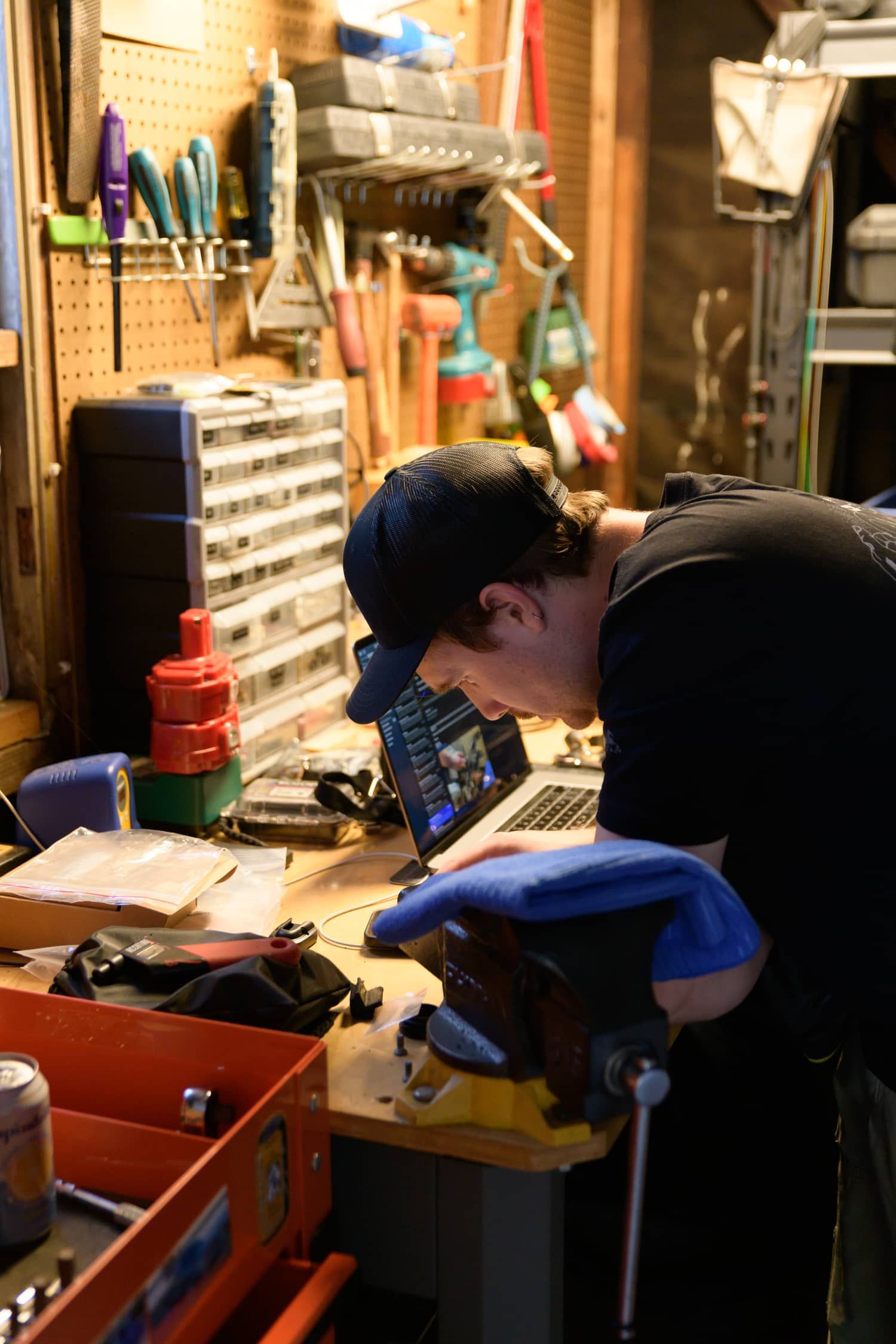 A mechanic in a black cap and gloves works at a cluttered workshop bench with a laptop open among various tools.