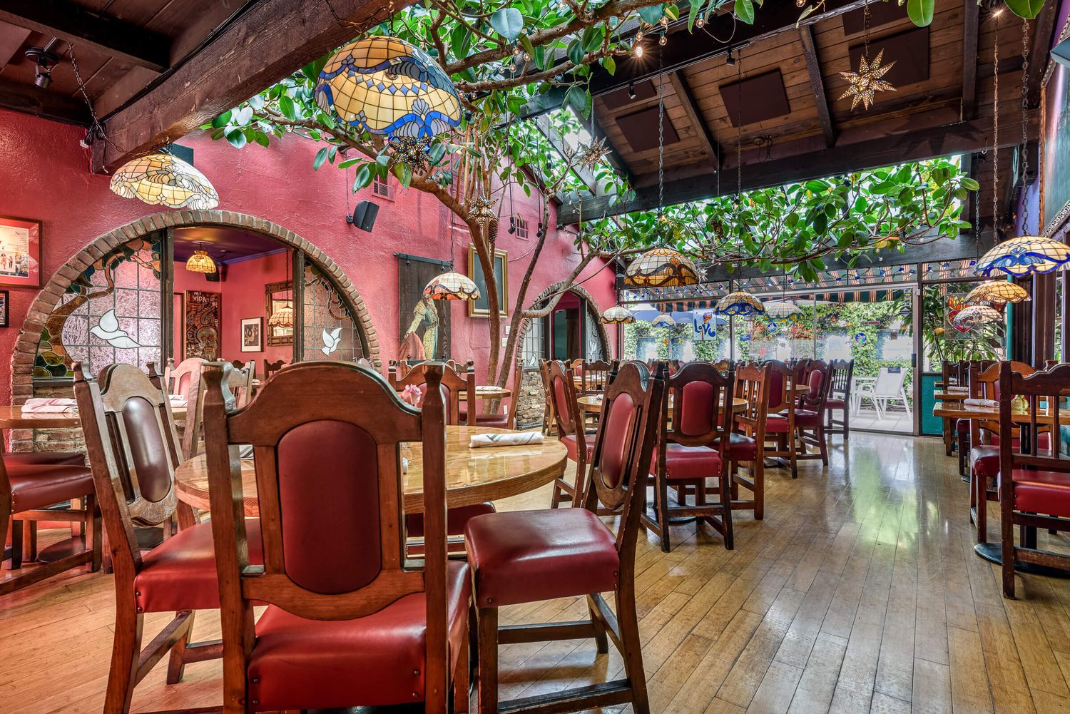 Colorful Mexican restaurant interior with red walls, wooden chairs, lush green vines, and decorative stained glass lamps overhead.