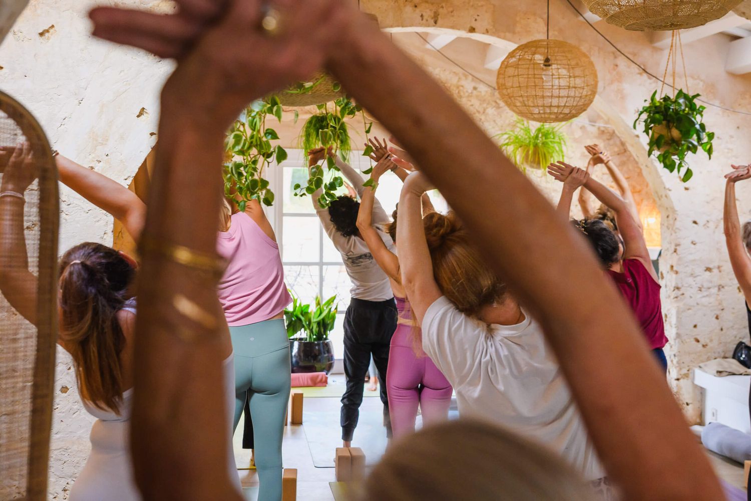 Group of women practicing yoga in a boho studio with hanging plants and rattan decor, arms raised overhead.