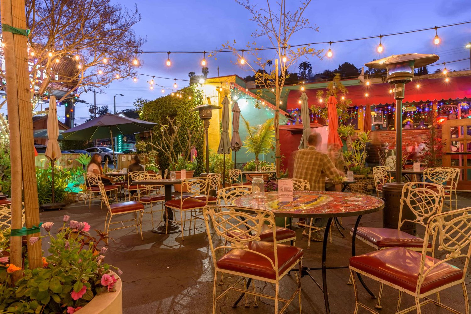 Colorful outdoor restaurant patio with string lights, ornate chairs, and lush greenery at twilight.