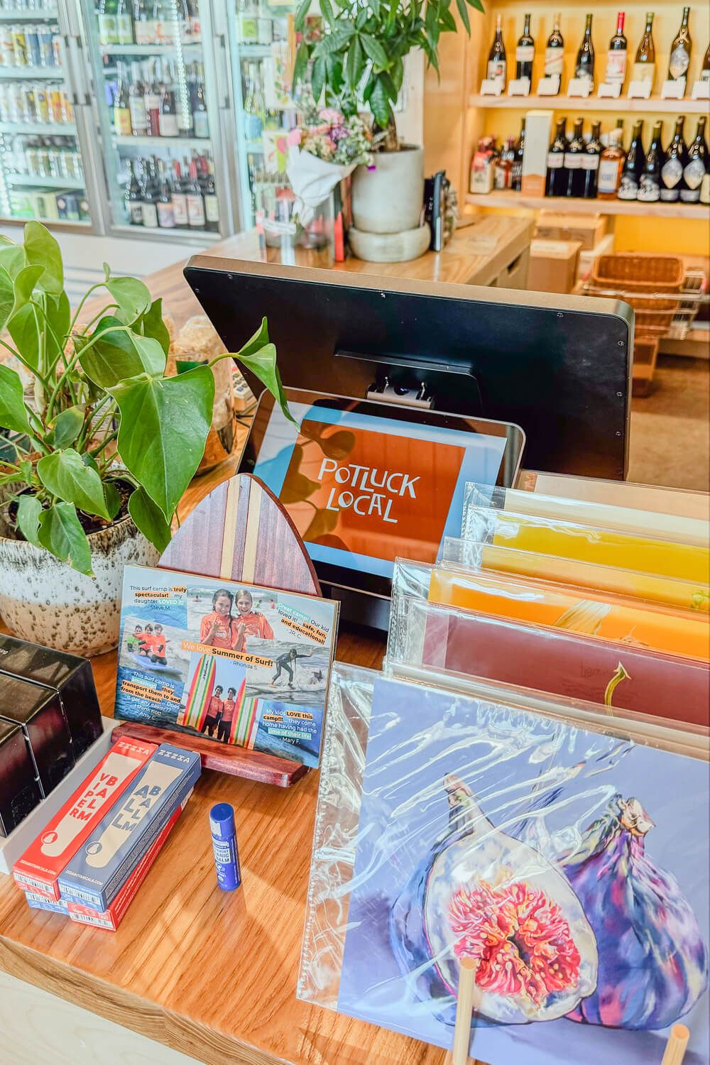 Potluck Local shop counter displaying colorful art prints, greeting cards, and merchandise with wine bottles on shelves in background.