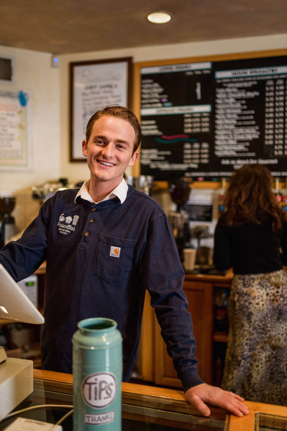 Smiling barista in navy Carhartt shirt stands at coffee shop counter with tips jar and menu board behind.