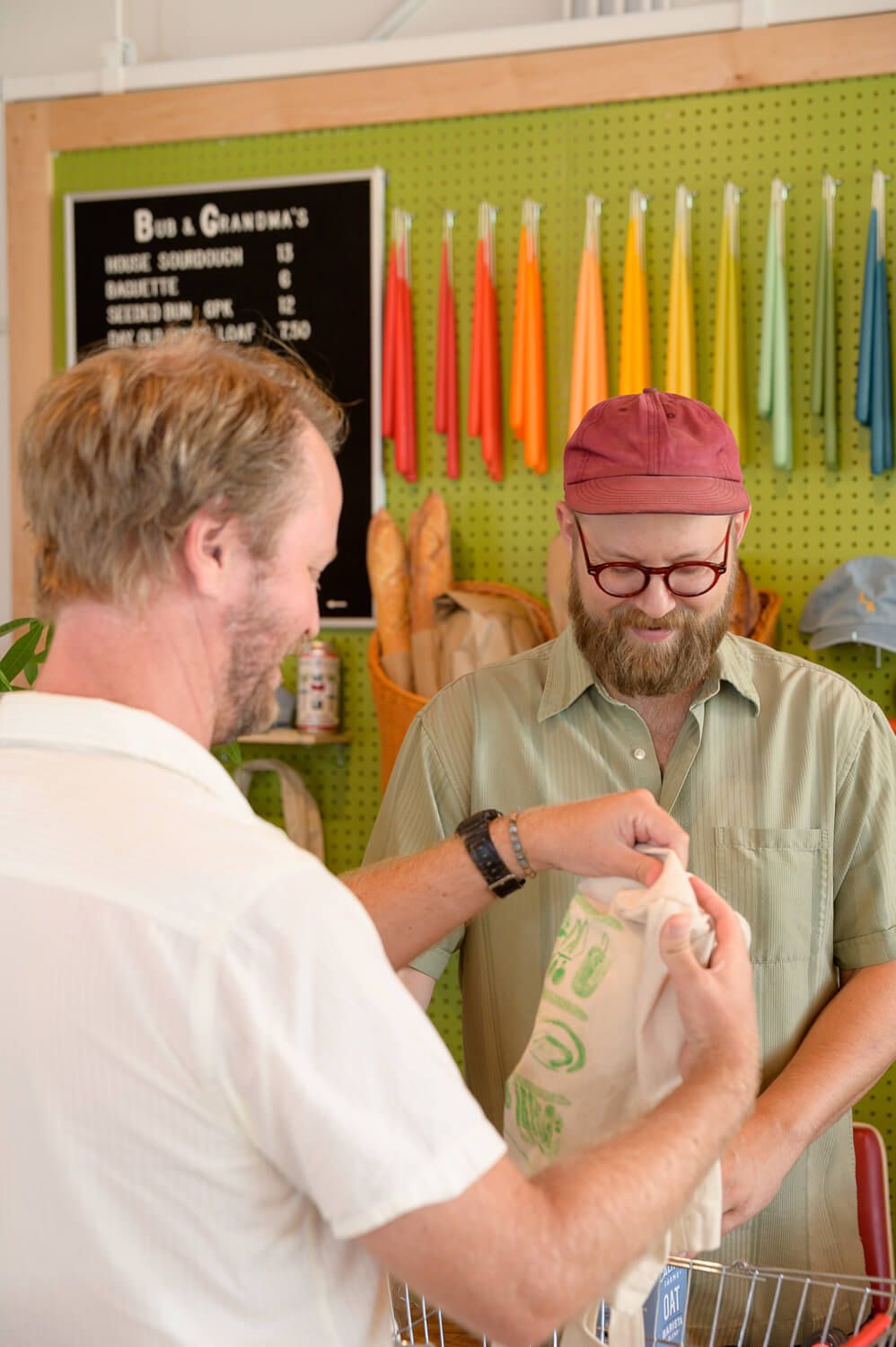 Two men at a bakery counter, one handing a bag of bread to a bearded man in a red cap and glasses.