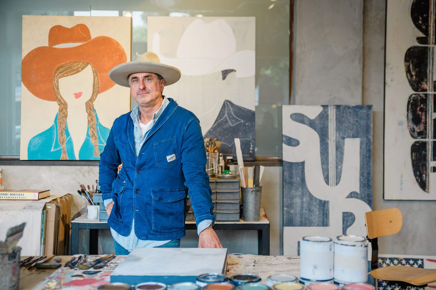 Artist stands in studio wearing blue jacket and wide-brim hat, surrounded by colorful paintings and art supplies.