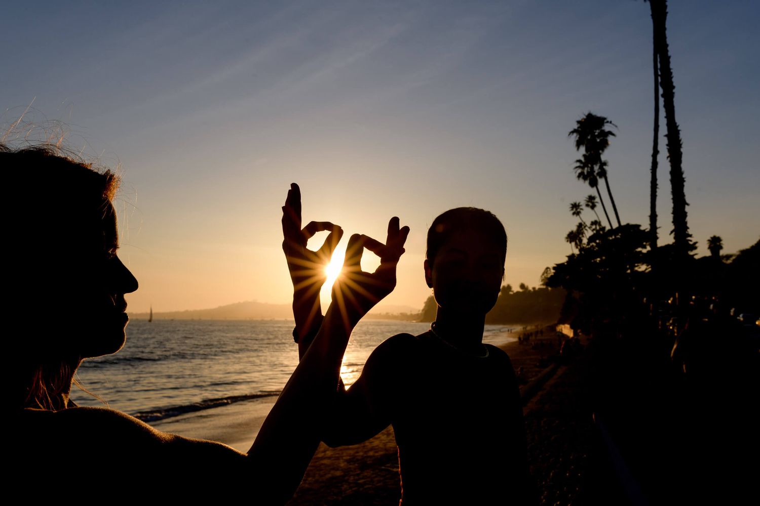 Silhouettes of two people making yoga hand gestures framing a golden sunset over the ocean with palm trees.
