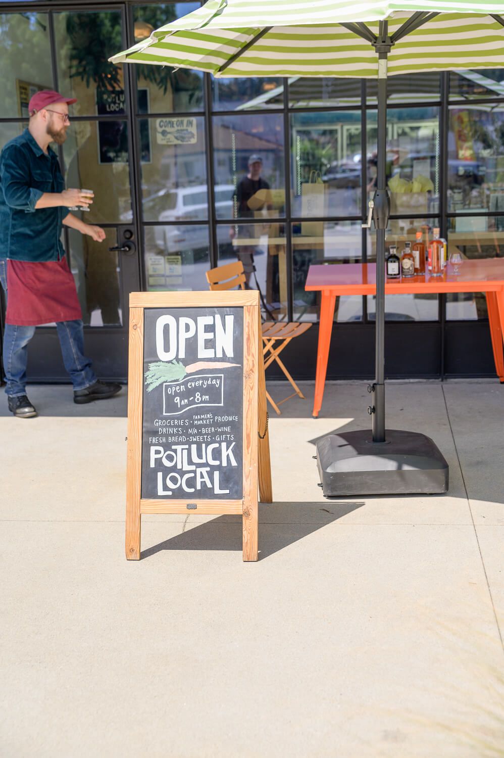 Outdoor chalkboard sign reading OPEN and Potluck Local outside a cafe with striped umbrella and server nearby.