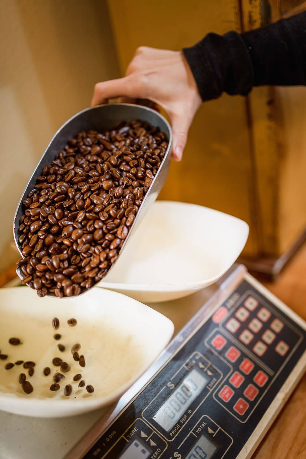 Hand pouring roasted coffee beans from metal scoop into white bowl on digital weighing scale.