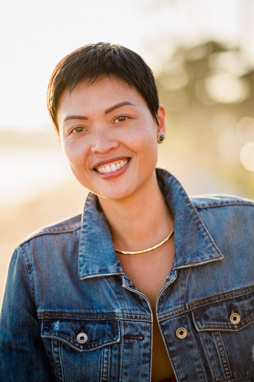 Smiling woman with short hair wearing a denim jacket and gold necklace outdoors in warm sunlight.