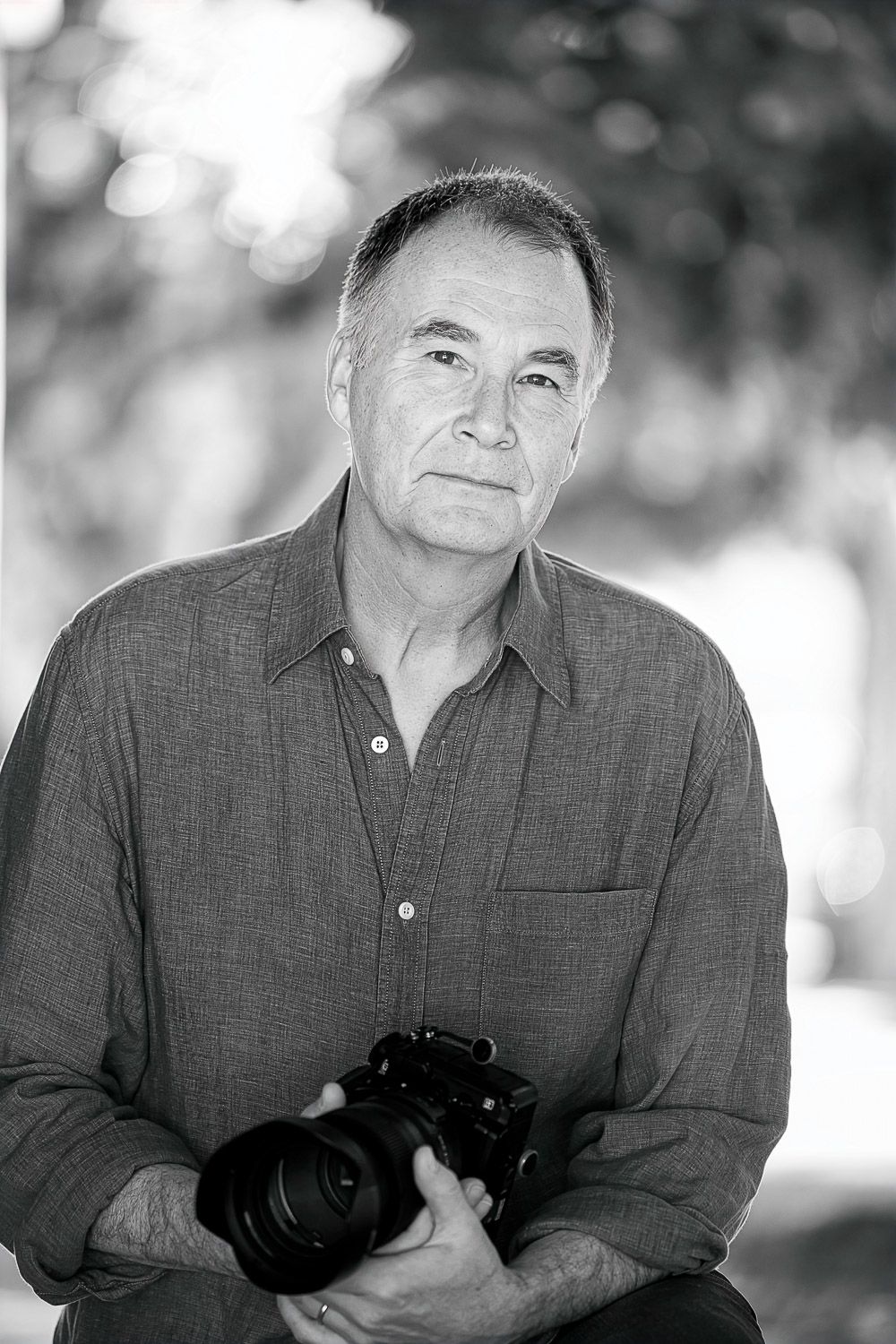 Black and white portrait of a mature man holding a DSLR camera outdoors with bokeh background.