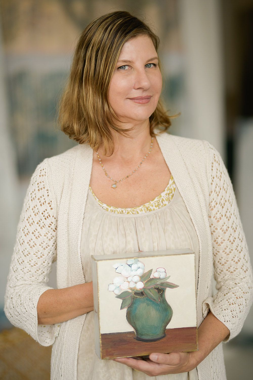Woman in cream cardigan holding a small painting of white flowers in a green vase on a wooden base.
