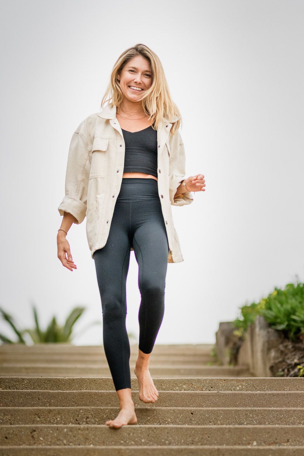 Smiling blonde woman in dark leggings, crop top, and cream jacket walking barefoot down outdoor steps.