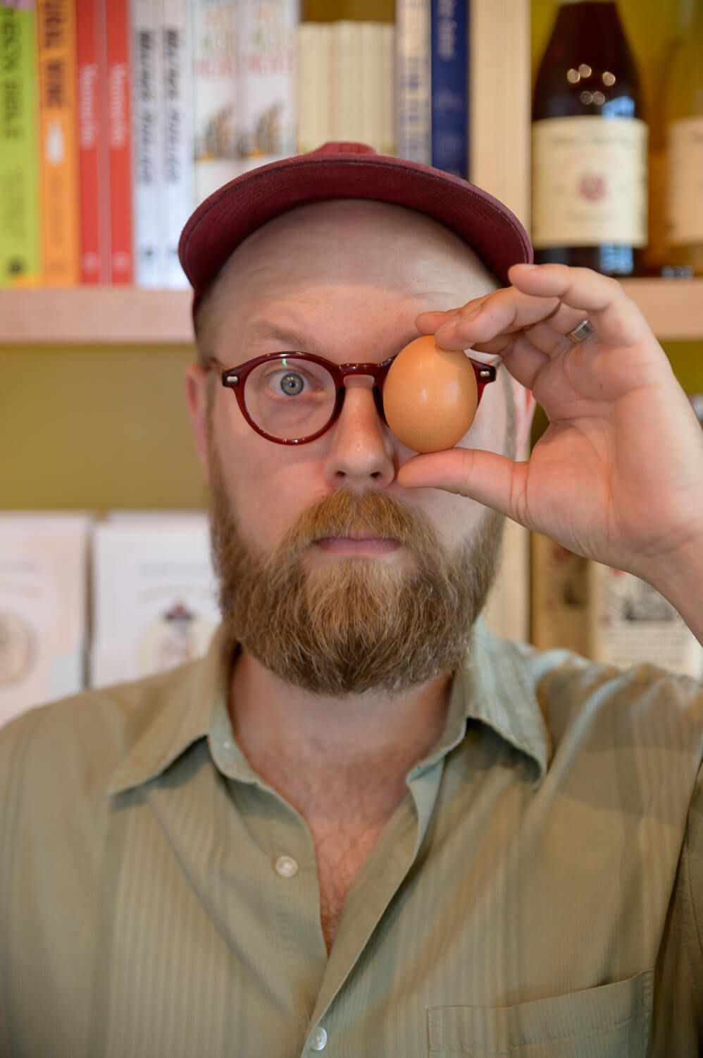Bearded man with glasses and red cap holds a brown egg in front of his eye in a bookshelf-lined room.
