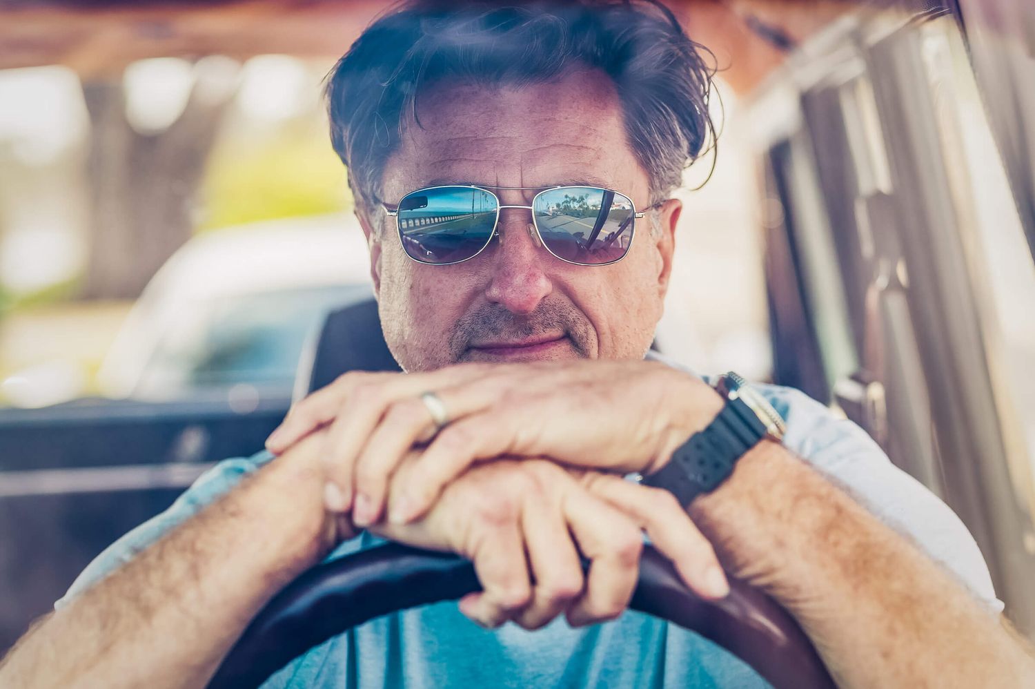 Middle-aged man with sunglasses smiling while leaning on car steering wheel, wearing a watch and blue shirt.