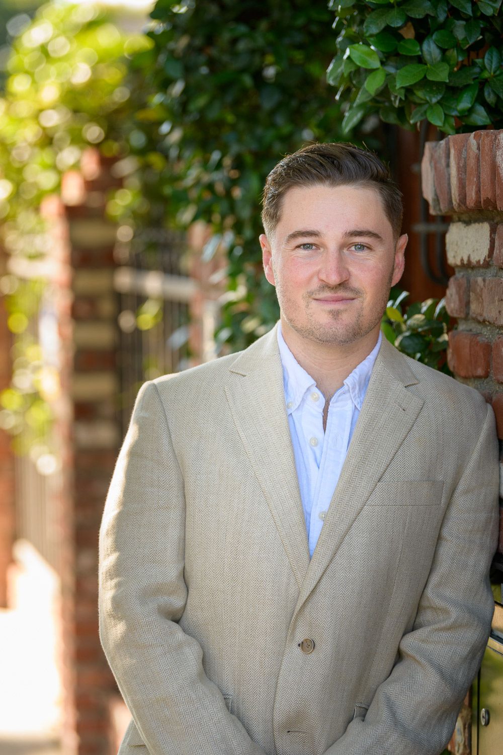 Professional man in beige suit jacket posing outdoors near brick wall with green ivy foliage in background.
