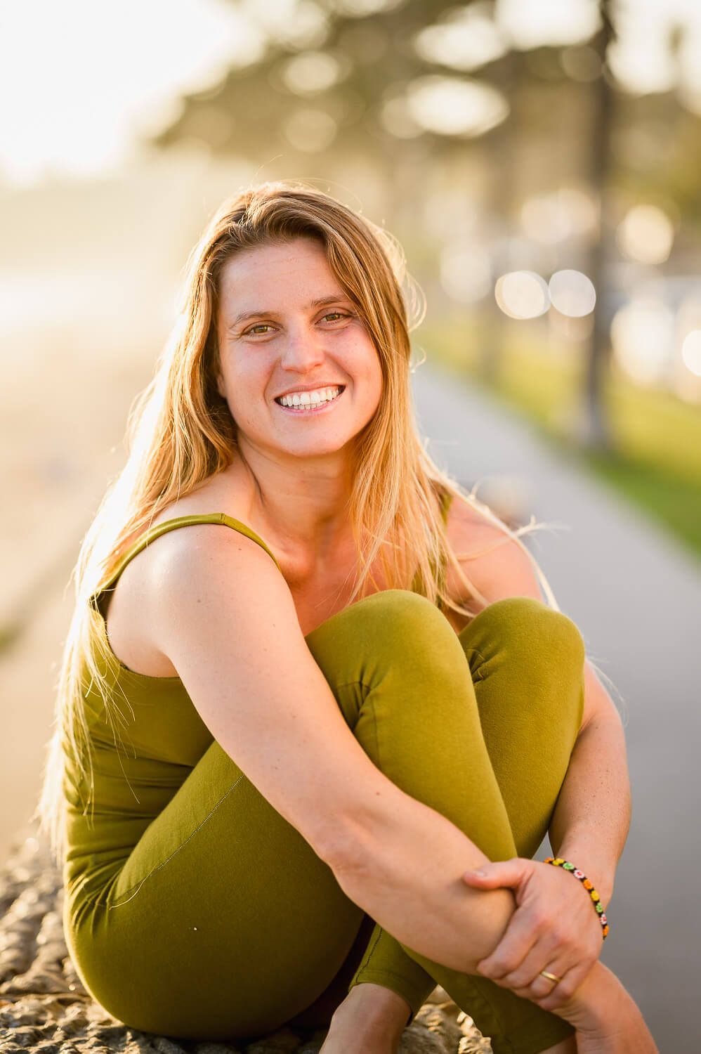 Smiling blonde woman in olive green outfit sitting outdoors on sunny day with blurred tree-lined path background.
