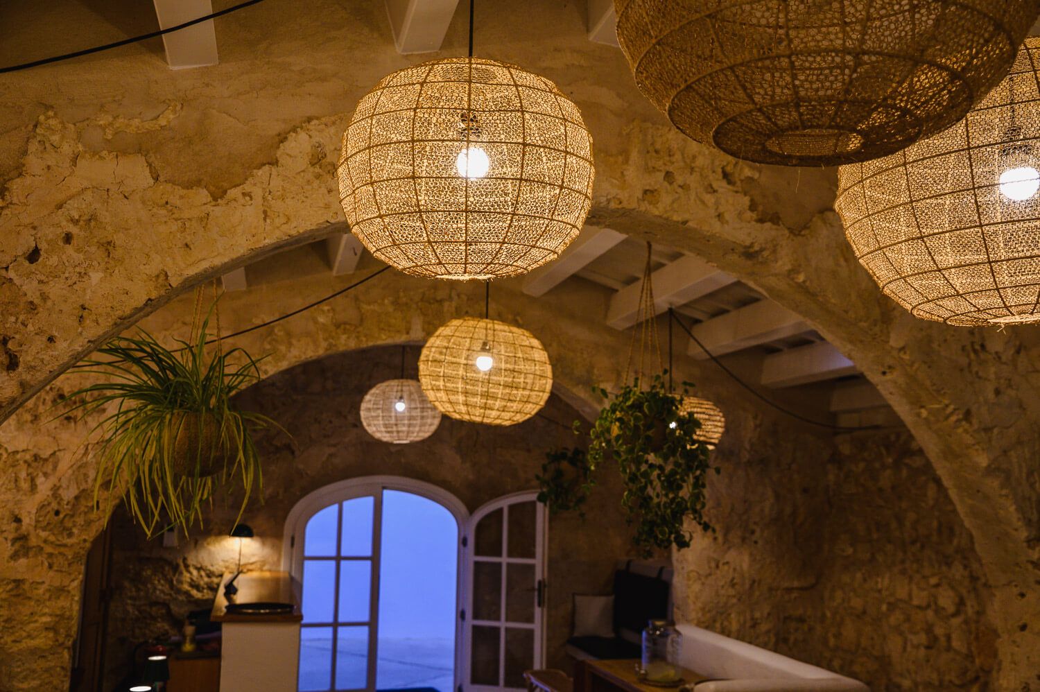 Wicker pendant lights hang from stone arched ceiling in a rustic Mediterranean restaurant with lush green plants.