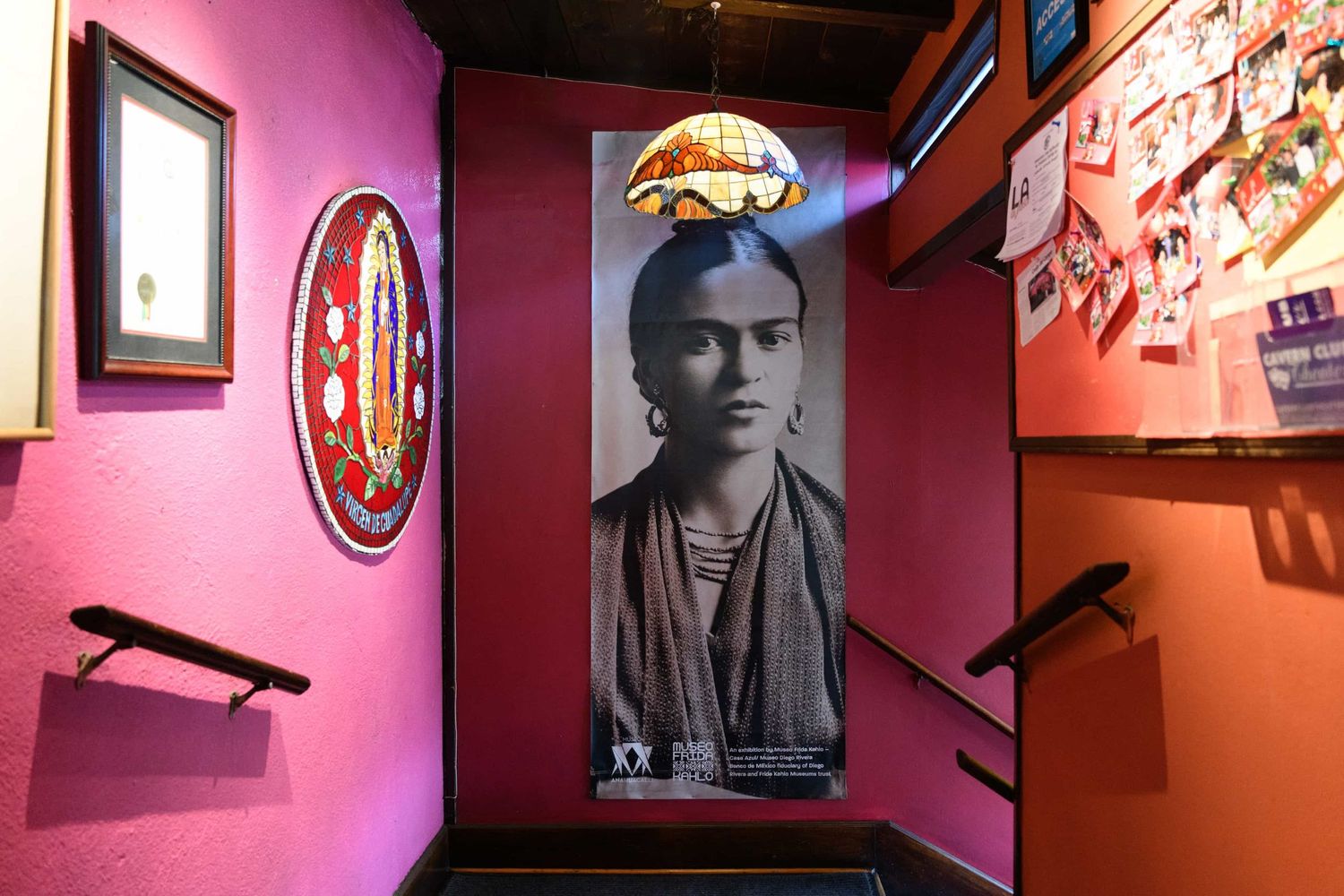 Colorful museum exhibit with a large Frida Kahlo portrait poster on pink walls, decorative art pieces, and ambient lighting.