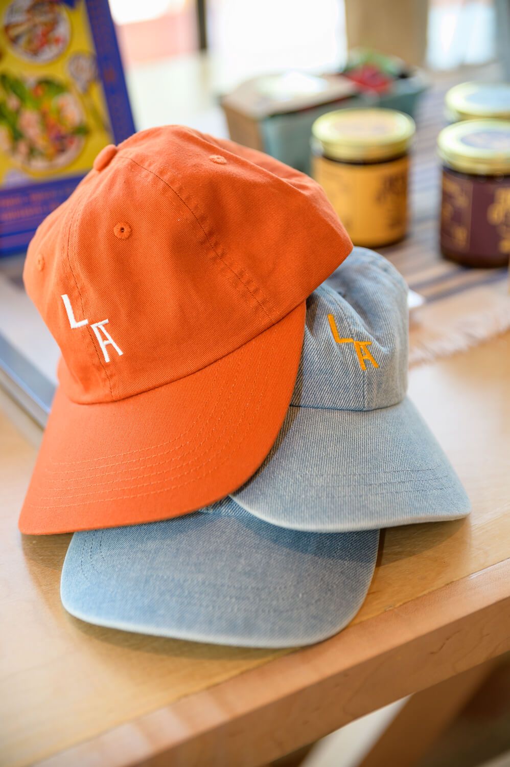 Two LA embroidered baseball caps in orange and light blue denim stacked on a wooden table.