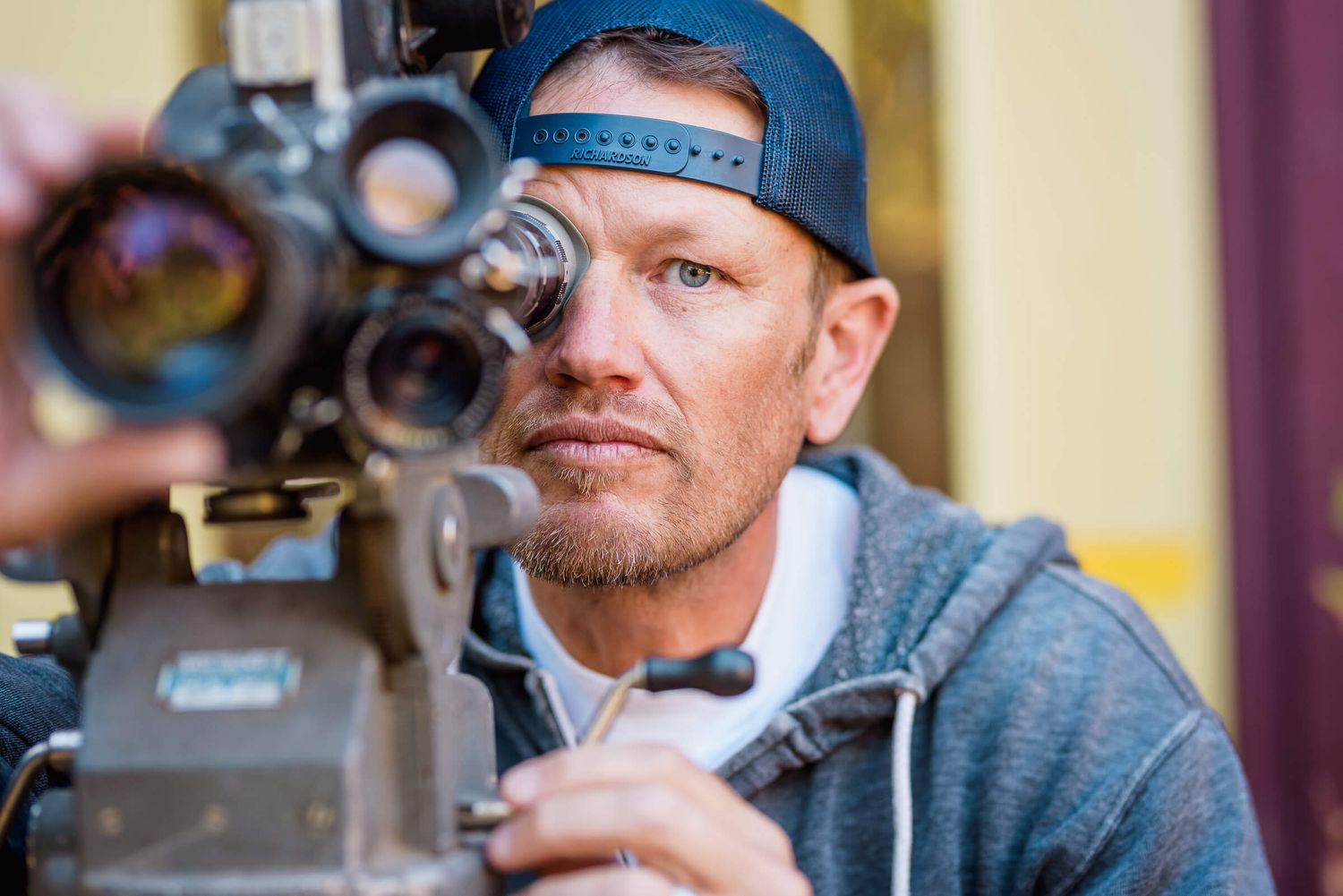 A focused male filmmaker looks through a vintage film camera while wearing a backwards cap and gray hoodie.
