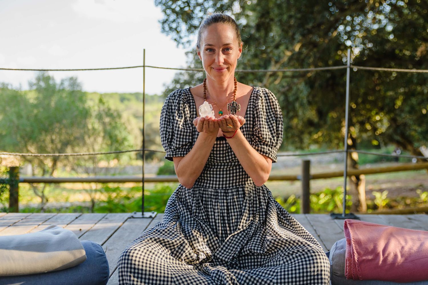 Woman in gingham dress sitting outdoors holding a crystal, surrounded by lush greenery and colorful cushions.