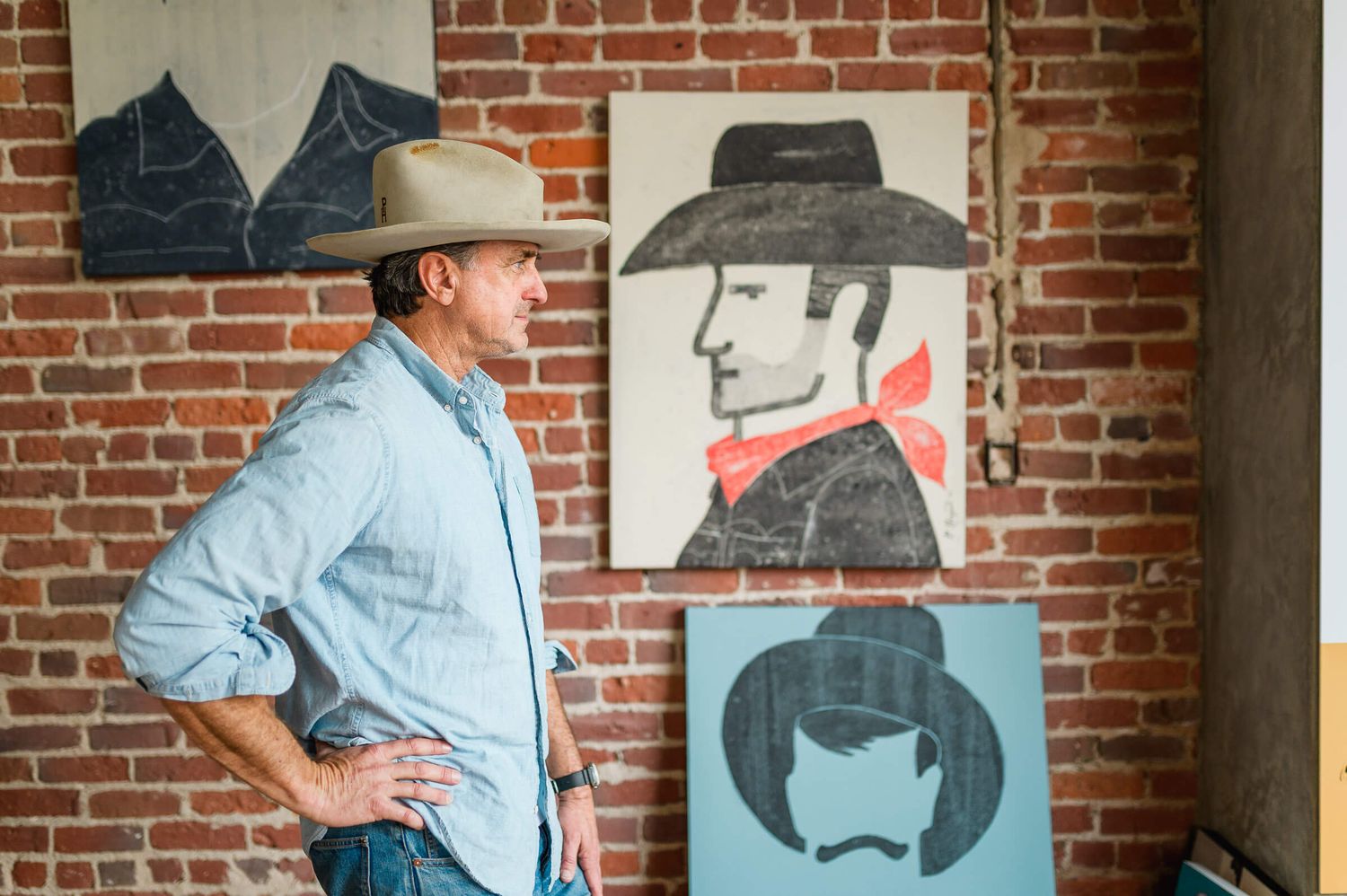 Man in cowboy hat stands beside Western-themed paintings on brick wall in rustic art studio.