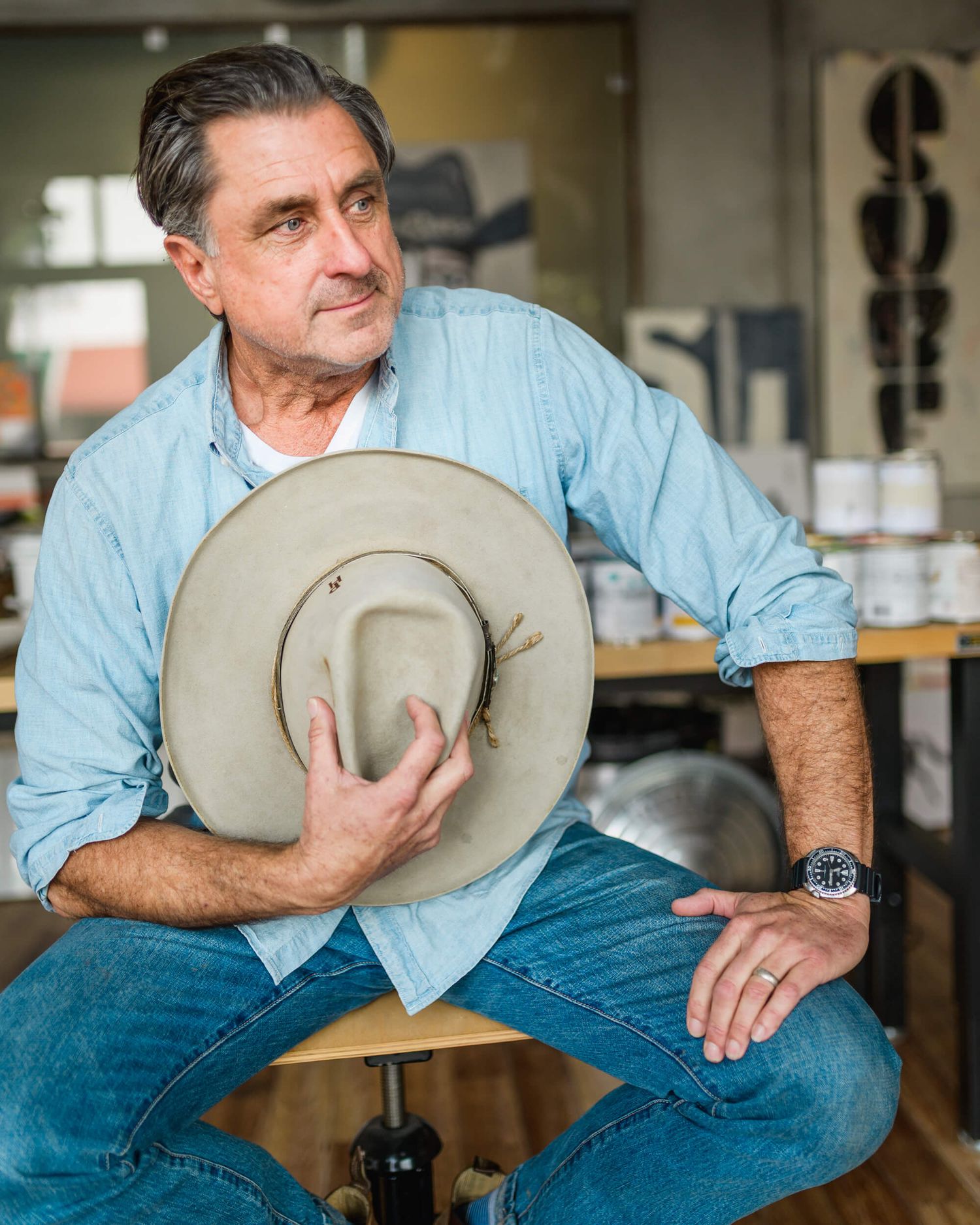 Middle-aged man in light blue shirt holding a tan cowboy hat while seated in a workshop studio.