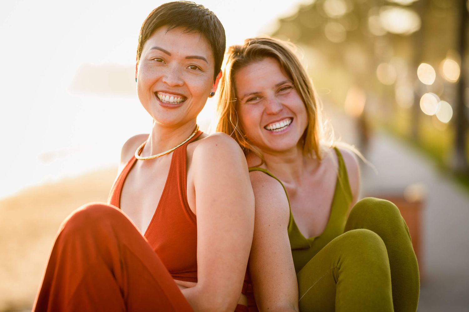 Two smiling women sitting together outdoors in warm golden sunlight, wearing red and green outfits.