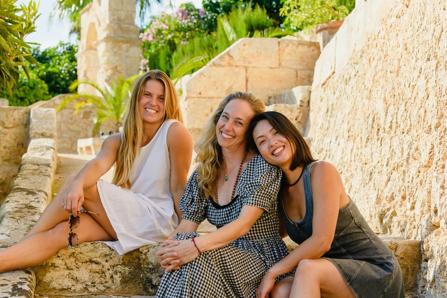 Three smiling women sitting together on stone steps surrounded by lush greenery and ancient architecture.