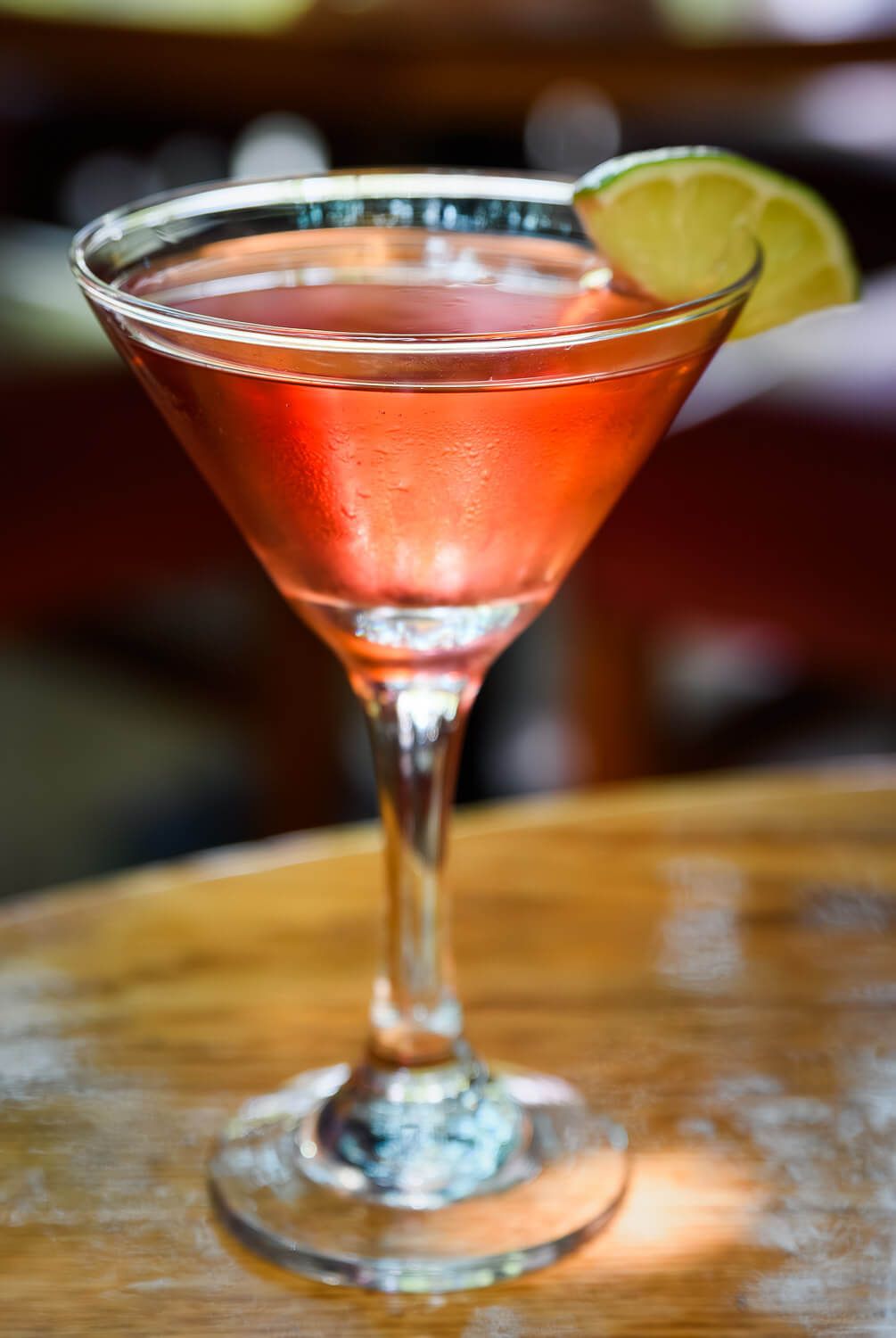 A vibrant pink cosmopolitan cocktail in a martini glass garnished with a lime wedge on a wooden bar.