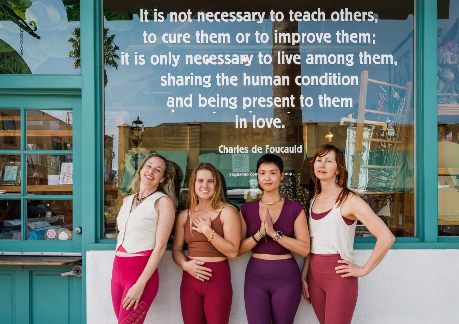 Four women in yoga attire pose outside a yoga studio with a Charles de Foucauld quote about living in love.
