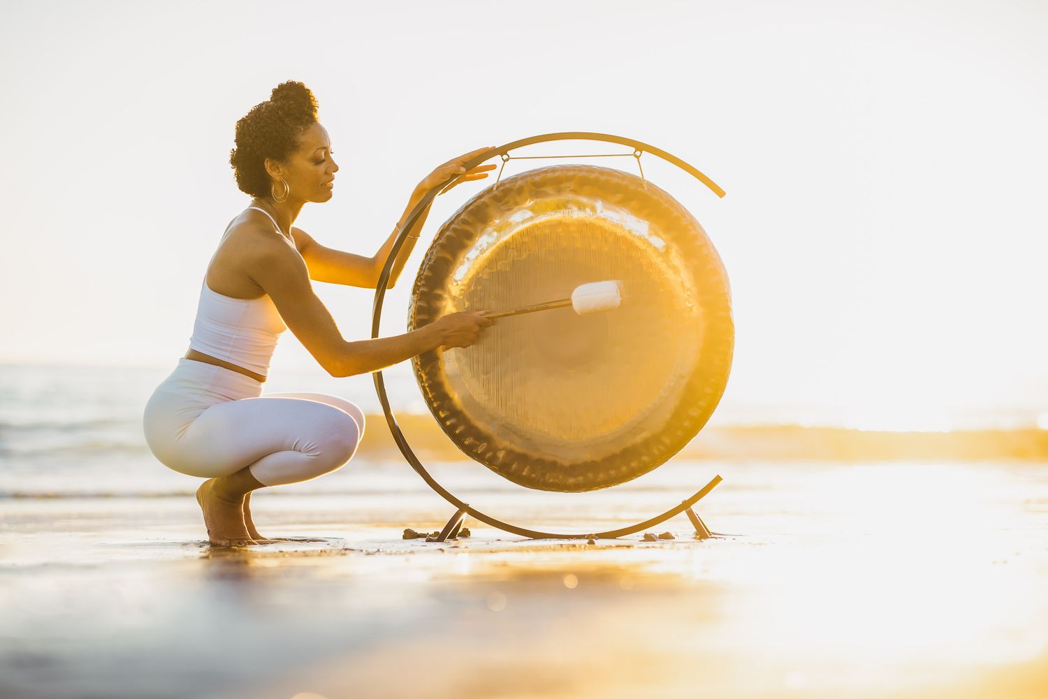 Woman in white activewear playing a large gong on a sunny beach at golden hour.