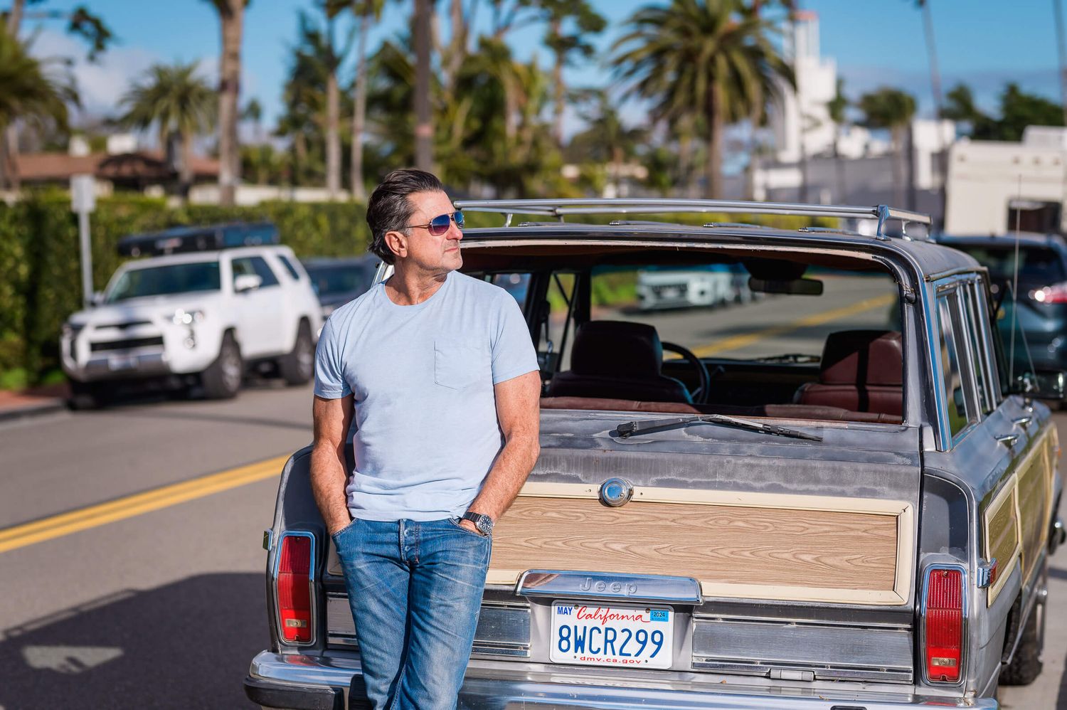 Man in sunglasses and jeans leaning against a vintage woody station wagon on a sunny California street.