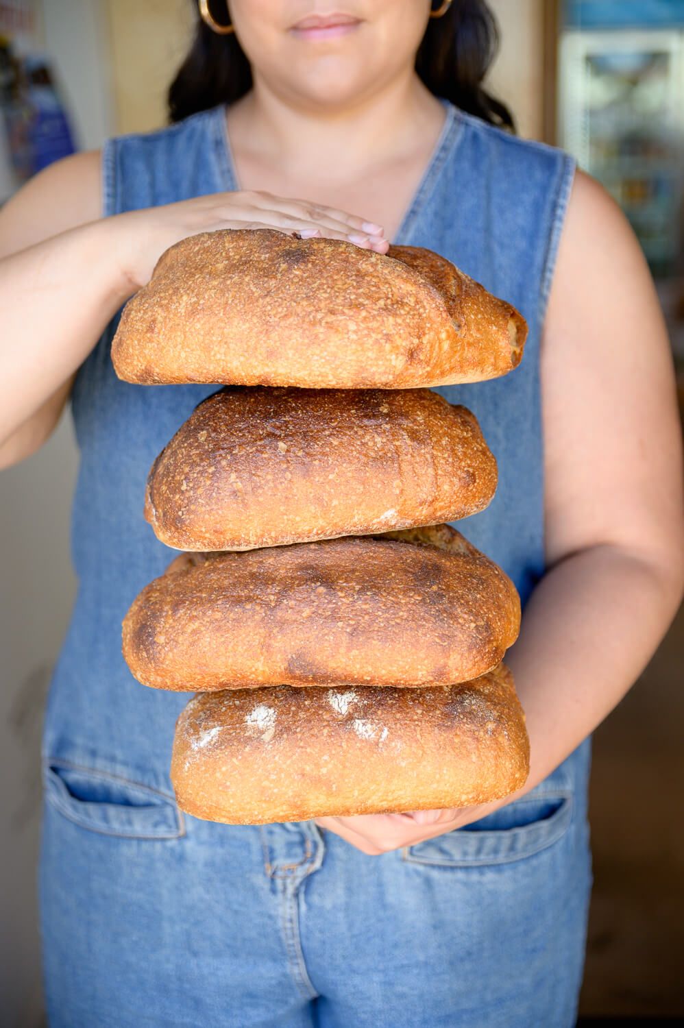 Person in denim overalls holding a stack of four freshly baked artisan bread loaves.