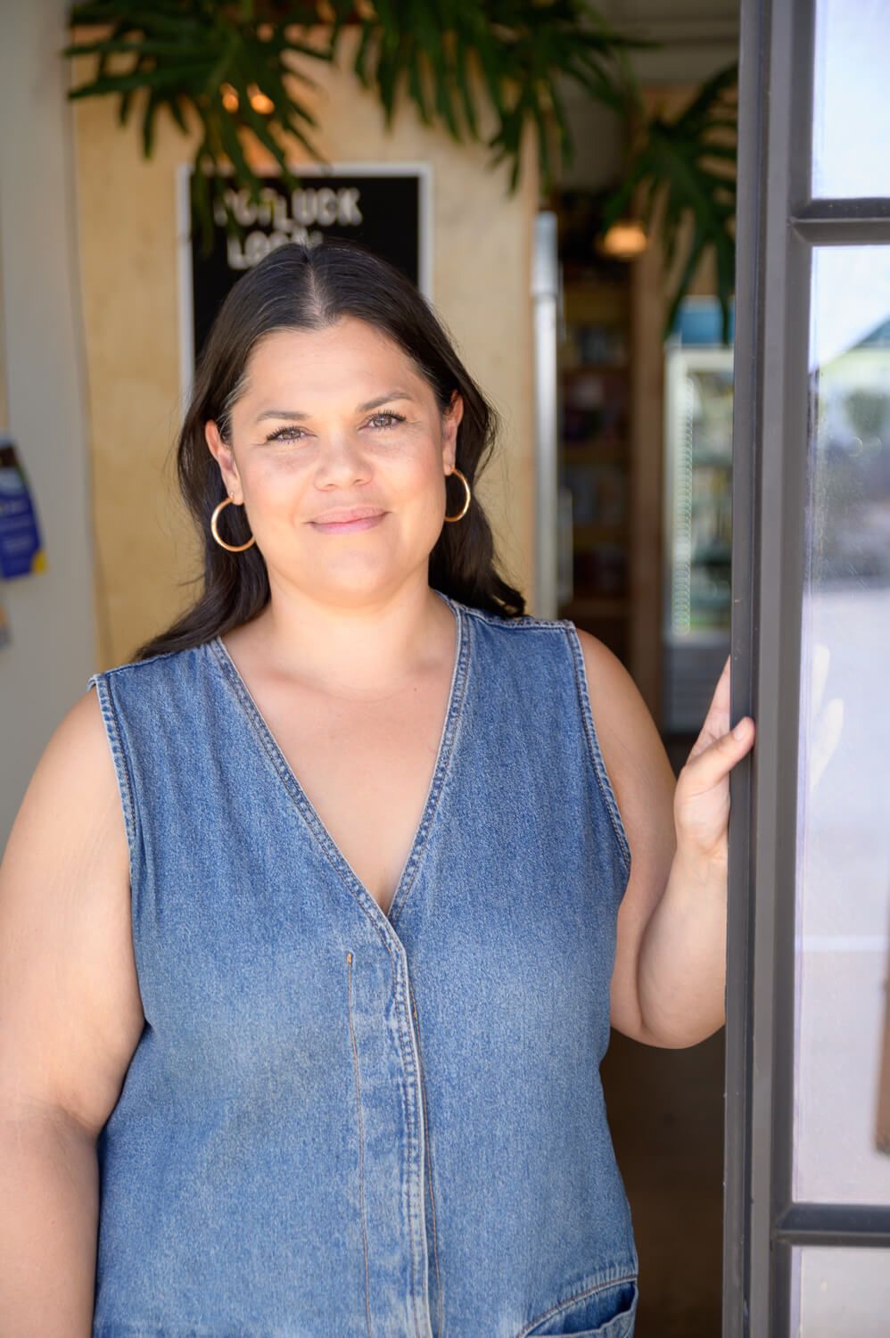 Woman in denim sleeveless dress with hoop earrings standing at a storefront door, smiling at camera.