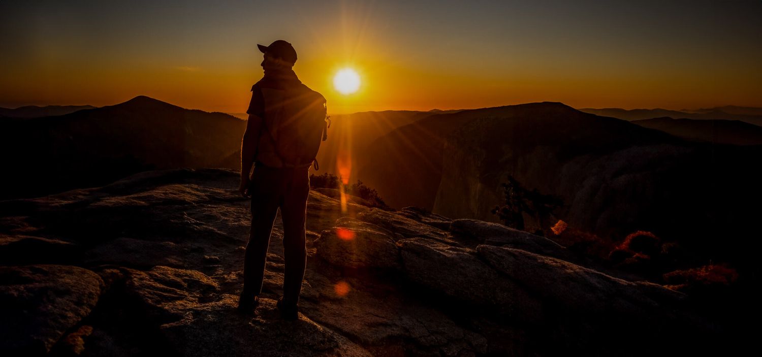 Hiker silhouetted against a stunning golden sunset on a rocky mountain summit with panoramic views.