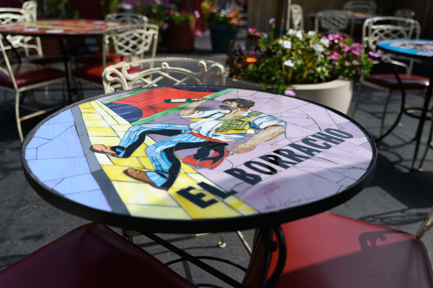 Round mosaic table with El Borracho comic art design at an outdoor cafe patio with colorful flowers.