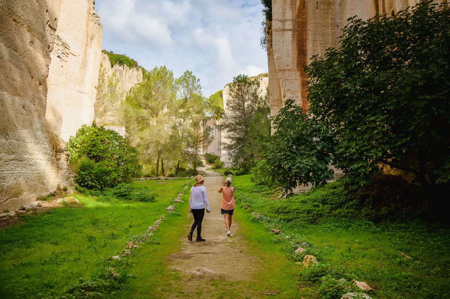 Two people walking on a scenic path between tall limestone cliffs and lush green trees in a natural canyon.