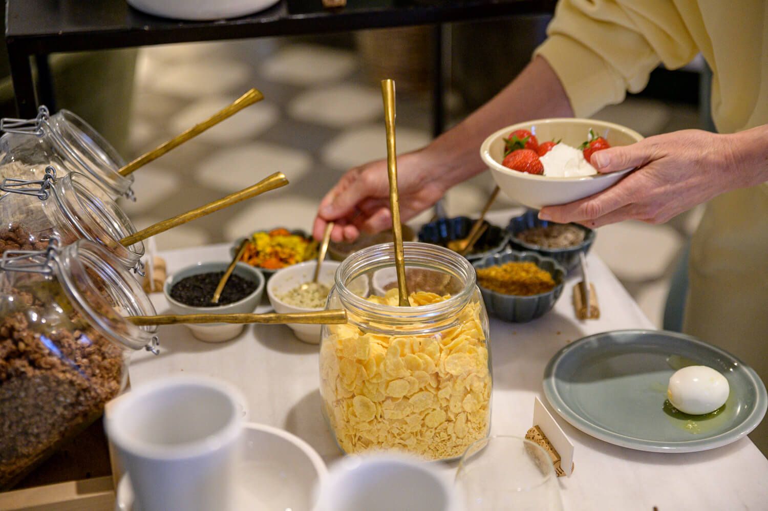A breakfast buffet spread with jars of cereals, fresh yogurt, and colorful toppings on a white counter.