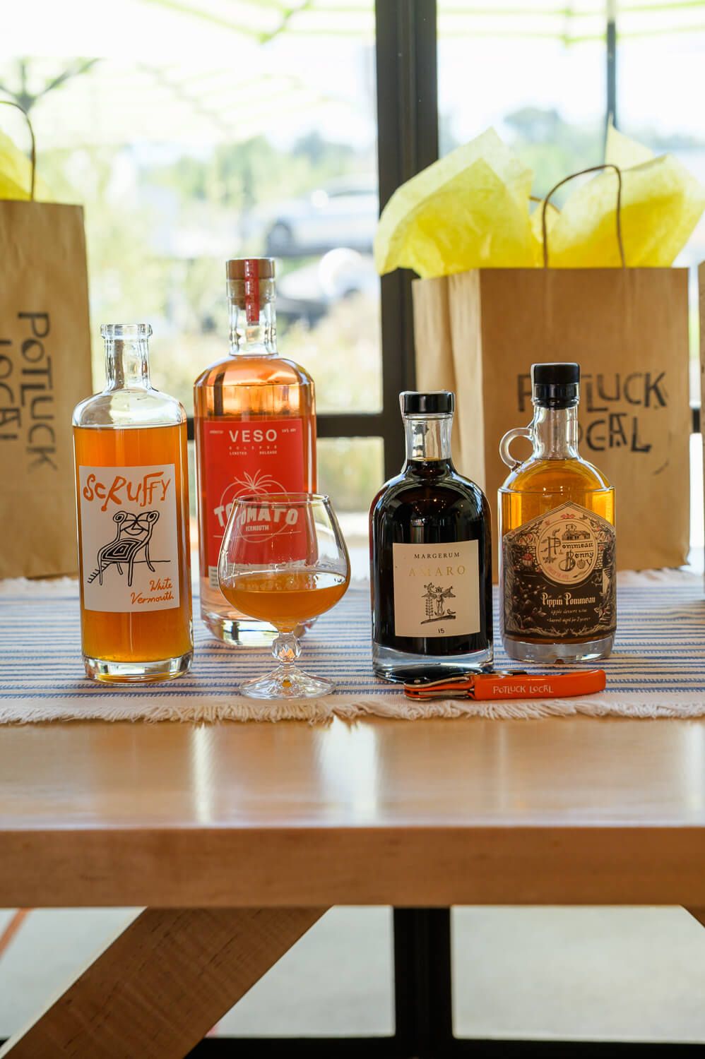 Assorted craft spirits bottles displayed on a wooden table with Potluck Local branded paper bags in background.