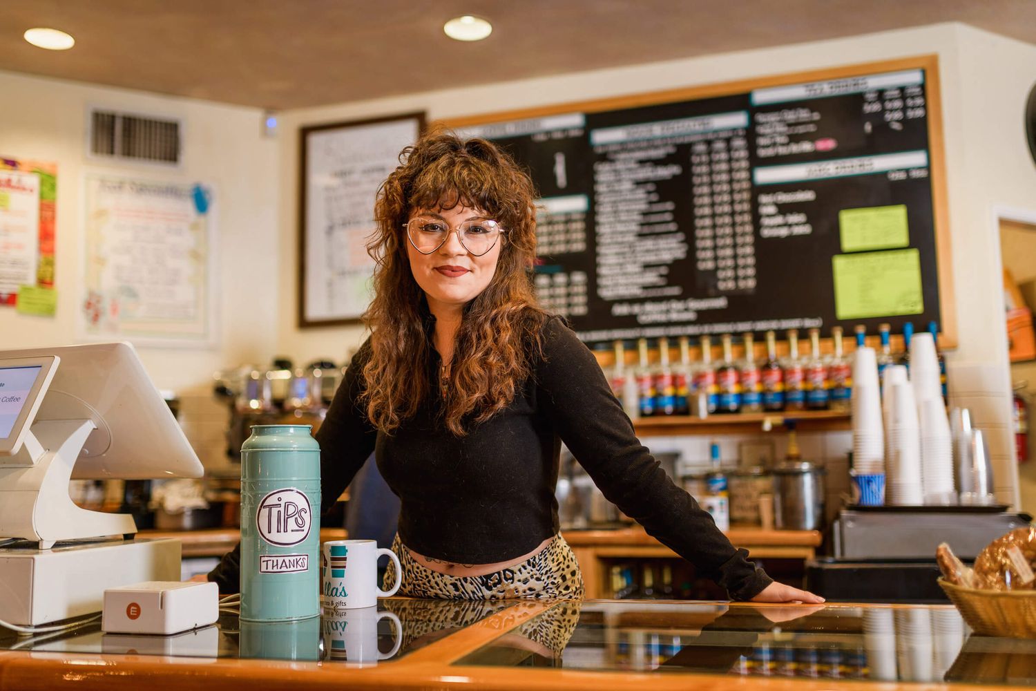 Young woman barista smiling behind coffee shop counter with menu board and teal water bottle.
