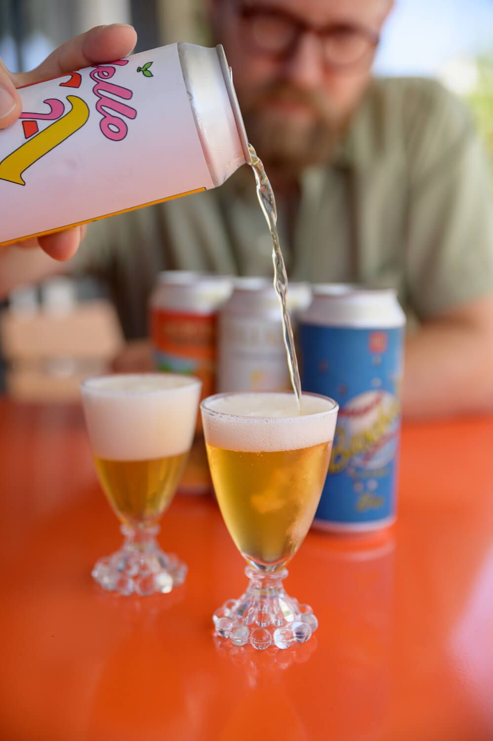 Person pouring craft beer from a pink can into glass goblets on a red table with blue beer cans in background.